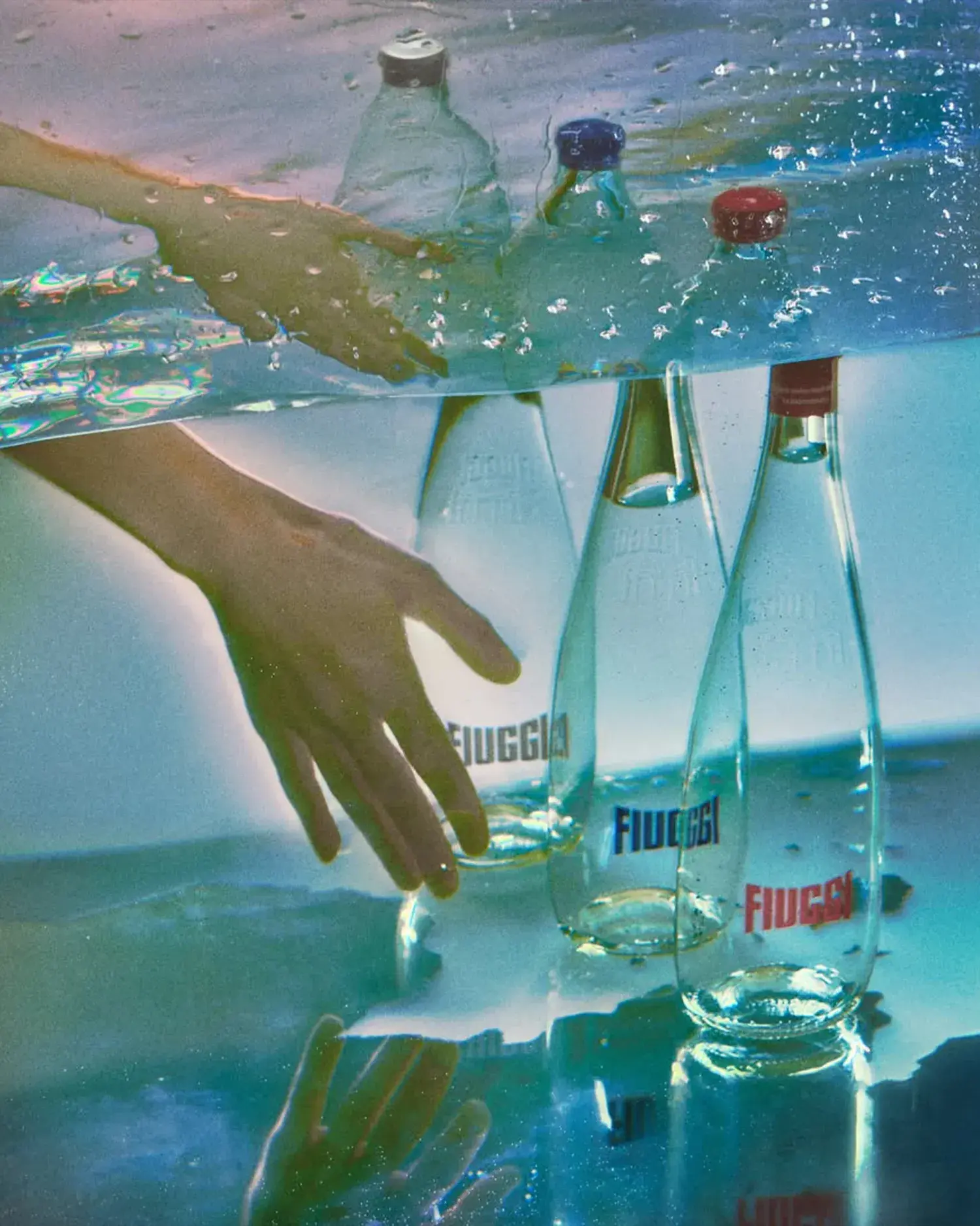 Three Acqua Fiuggi Luxury glass bottles with grey, blue and red caps submerged underwater, with a hand reaching towards them