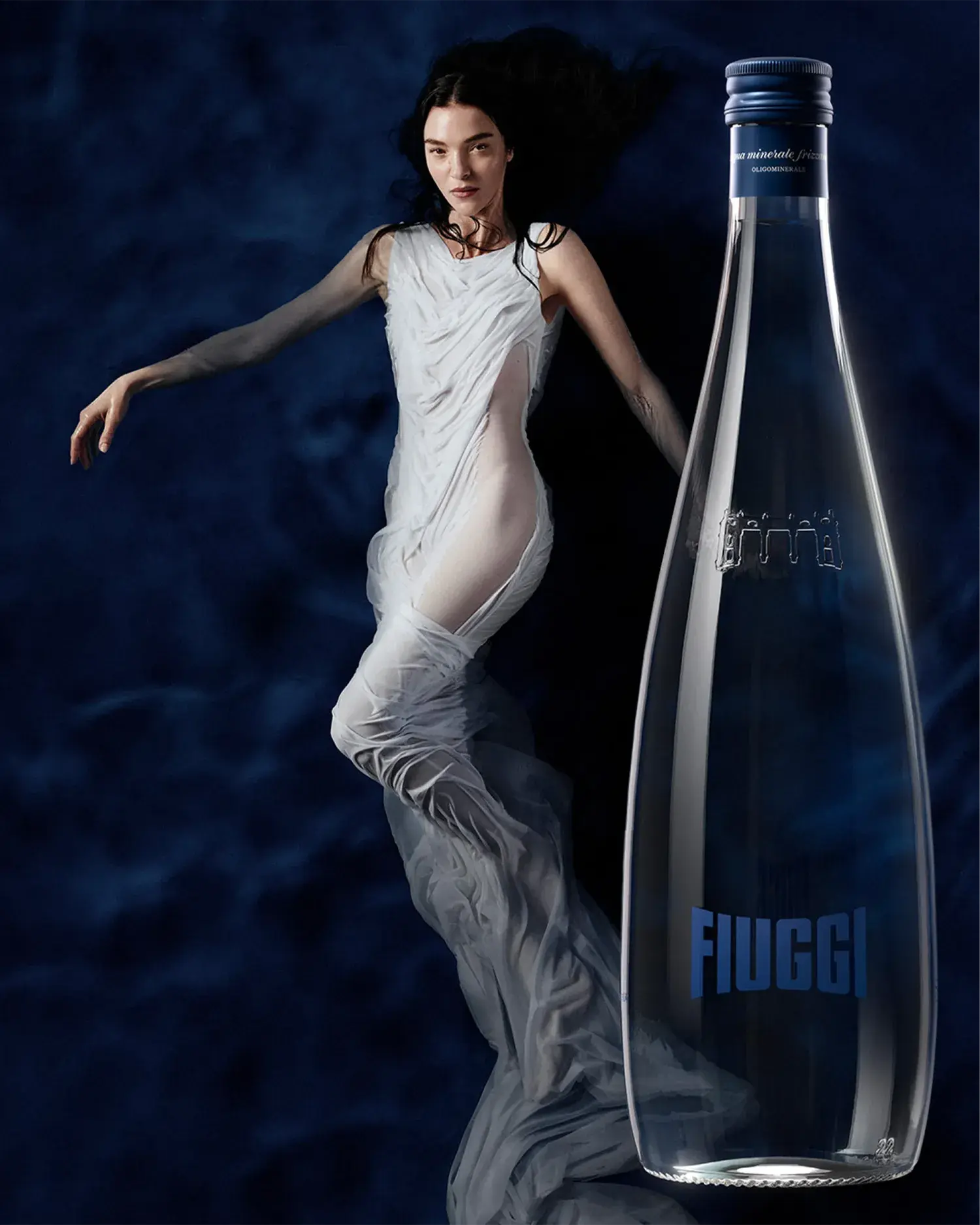 Model in a white draping dress next to an Acqua Fiuggi Luxury sparkling glass bottle with blue cap